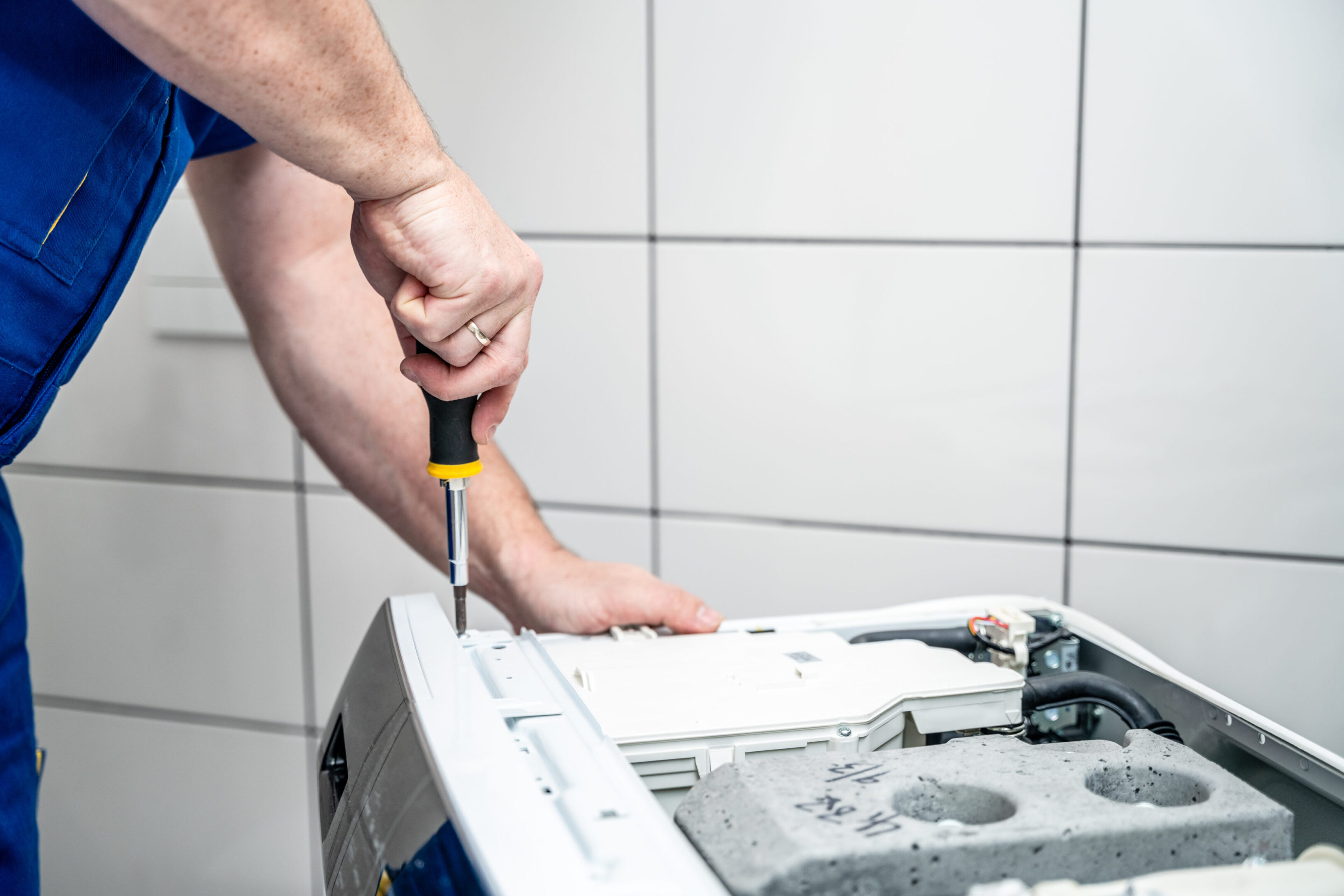 Repairman using a screwdriver disassembles a washing machine for repair Person in blue overalls using a screwdriver to repair or assemble a washing machine in a tiled room.